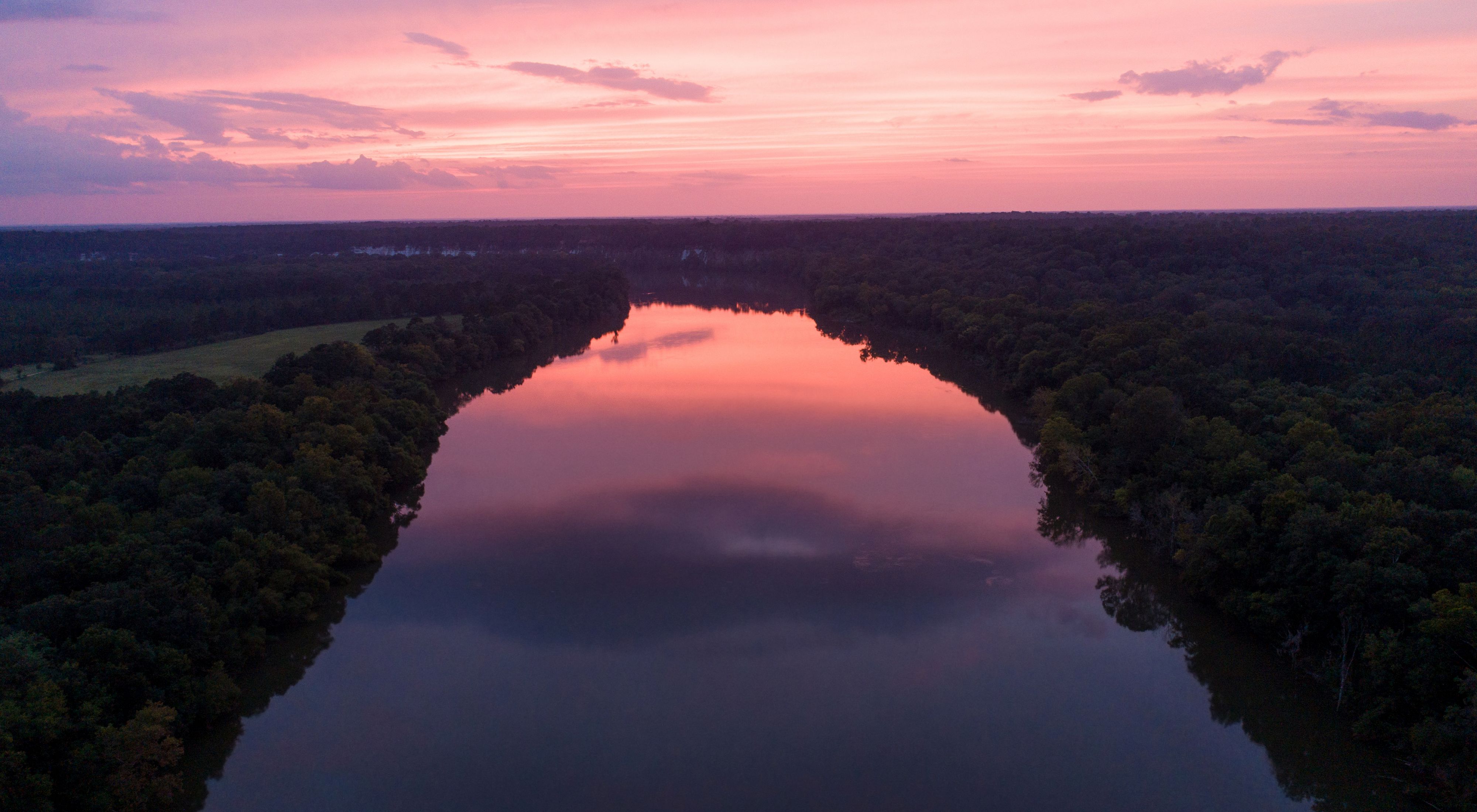 Sun sets over the Alabama River with pink and purple hues bouncing off the wide river in the center flanked by tall trees.