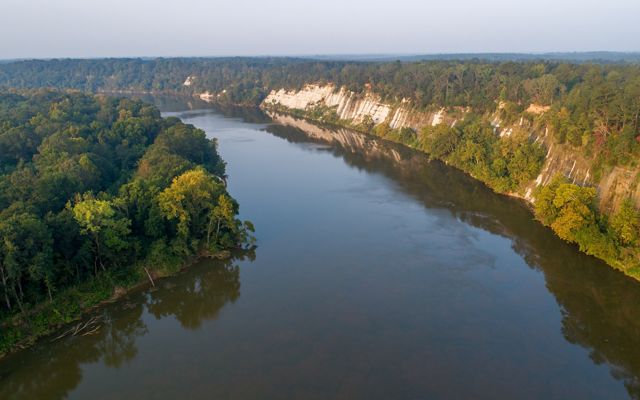 Tall rock formations known as bluffs reach up on one side of a river while the other side is flat.