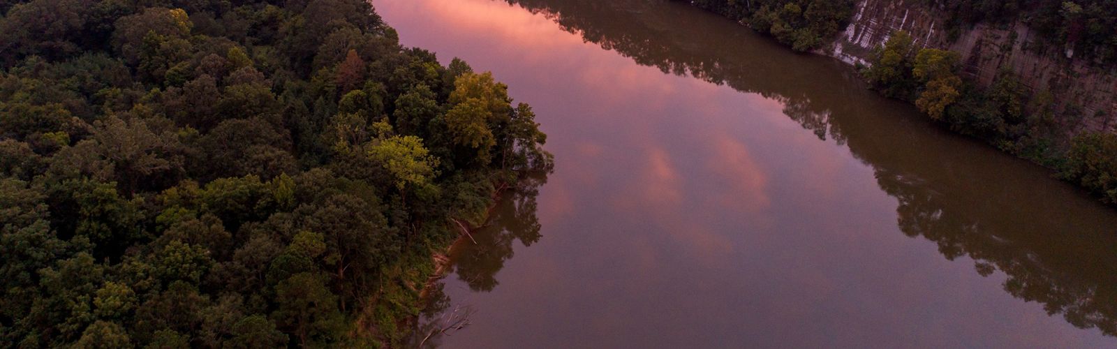 A river bordered by tall rocky bluffs on one side and dense forest on the other reflects the sunset in purple and pink hues.