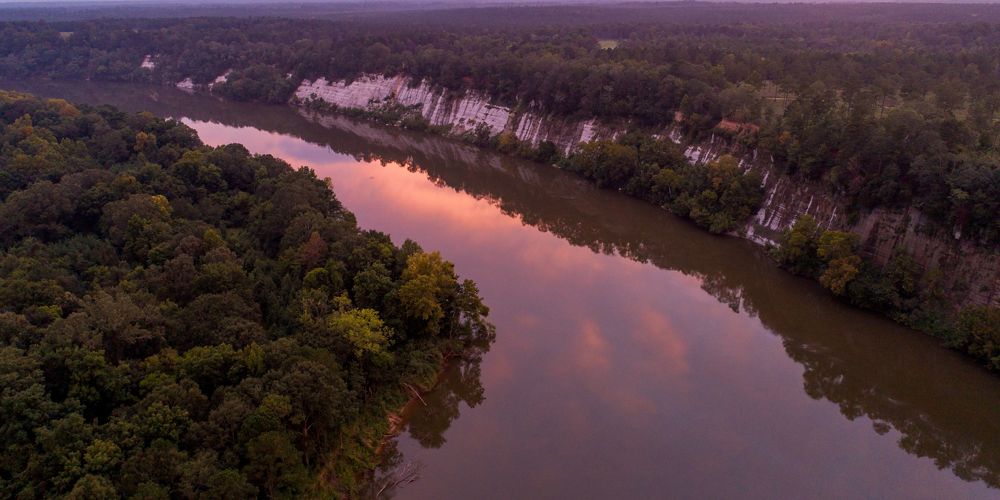A river bordered by tall rocky bluffs on one side and dense forest on the other reflects the sunset in purple and pink hues.
