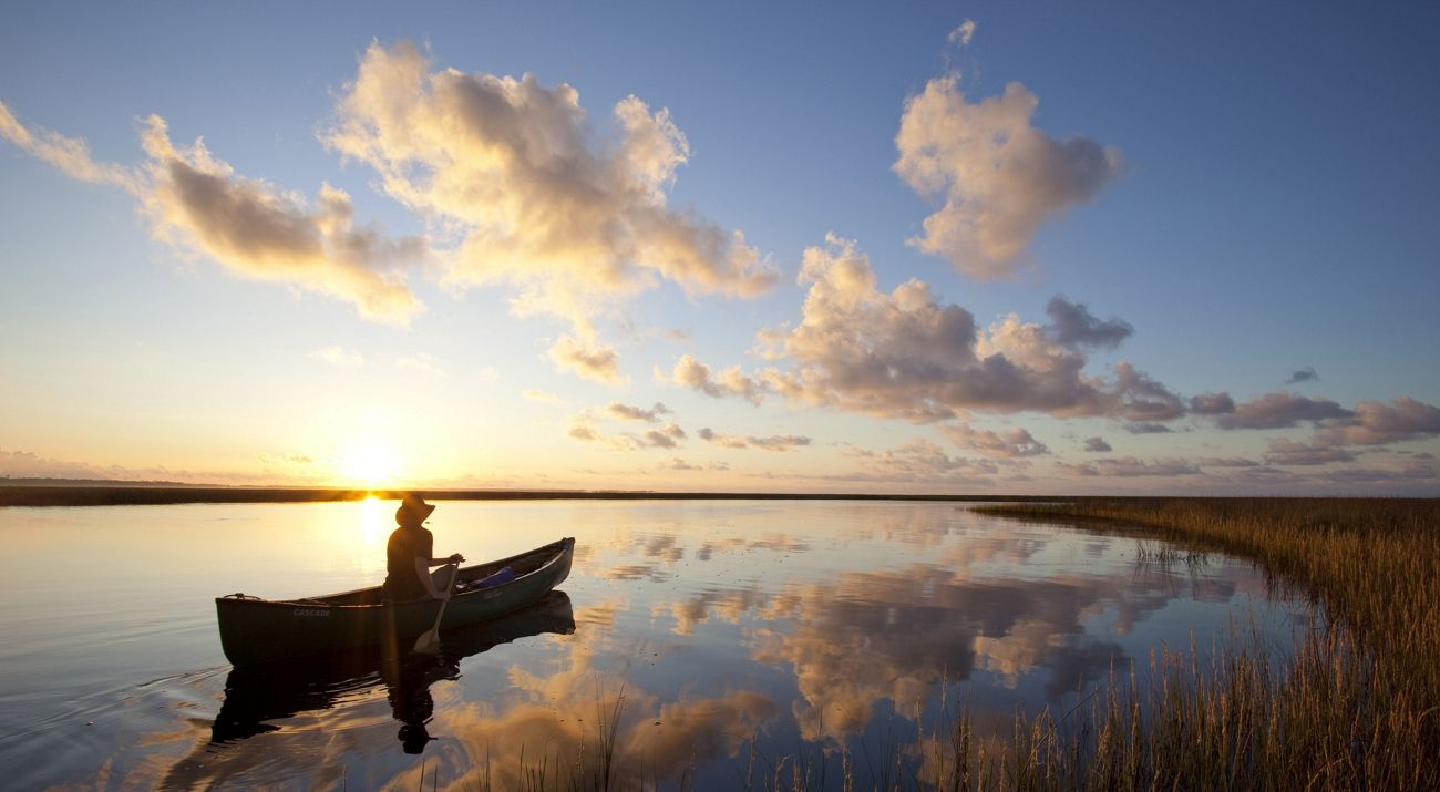 A person in a canoe pauses on calm waters as the sun sets.