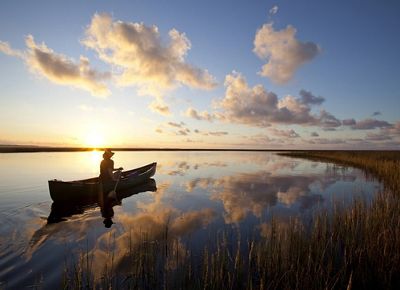 A person paddles a canoe across placid waters as the sun sets on the horizon.
