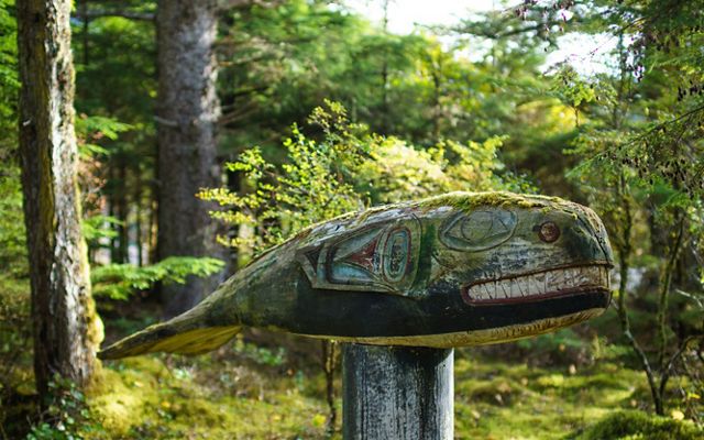Closeup of a wooden totem of a whale mounted atop a wooden post in a forest.