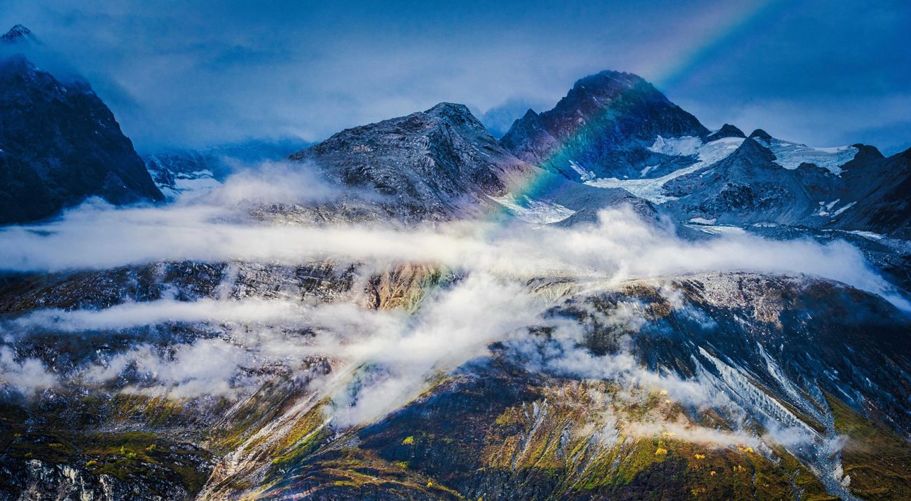 The peaks of a rugged mountain range are wreathed with long, white clouds and a rainbow arcs across the vista.
