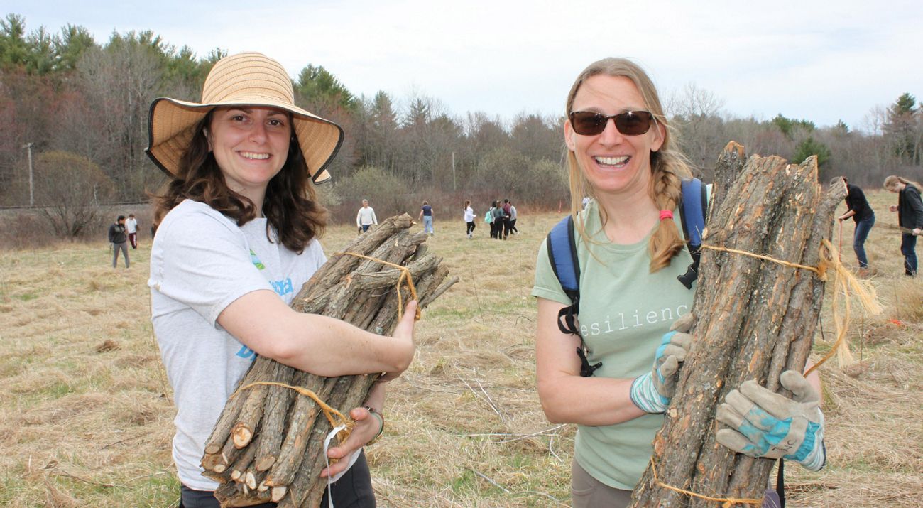 Two women, one wearing a hat and the other sunglasses, smile and hold bundles of wood in a field while people work on planting in the background.