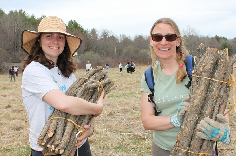 Two smiling woman stand together during a volunteer event. Each of them hold burlap wrapped bundles of young tree saplings.