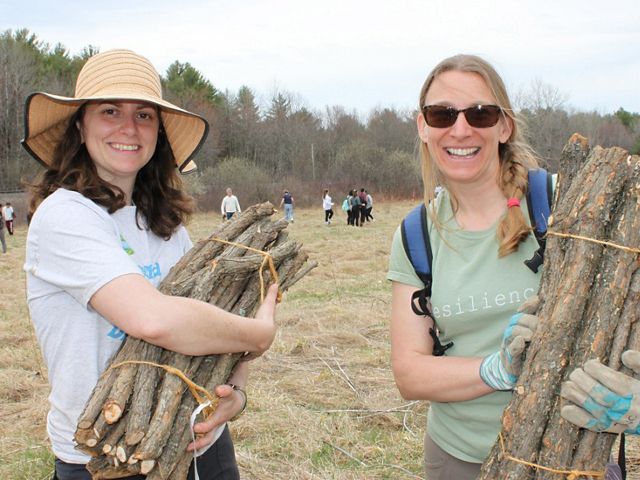 Two smiling woman stand together during a volunteer event. Each of them hold burlap wrapped bundles of young tree saplings.