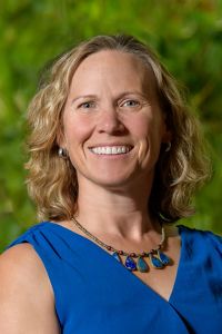 Headshot of a woman with a blue dress and necklaces and blond short hair smiling at the camera.