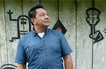 Um homem de camisa azul sorri olhando para o horizonte enquanto está em frente a uma estrutura de madeira pintada com iconografia indígena tradicional.
