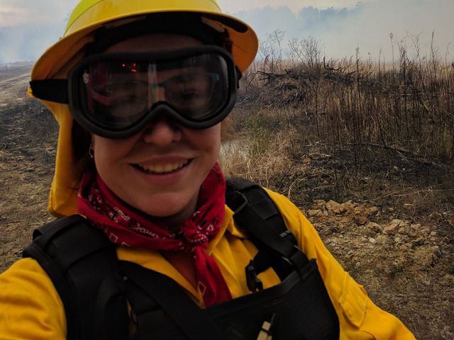 A woman wearing yellow fire retardant gear and thick goggles takes a selfie during a controlled burn. She is standing in front of an open scrubby piece of land that has not yet been ignited.