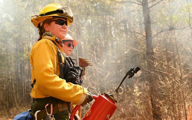 Andi Clinton holds a red metal drip torch. Smoke rises behind her following the ignition of a fire line at the WTREX fire learning exchange.