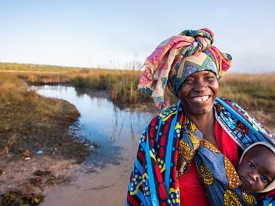 Una mujer sonriente carga a su niño en un harnés de telas coloridas frente a un río.