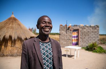 Retrato de líder Ferando Kufuna. Un hombre sonriente de pie frente a construcciones tradicionales.