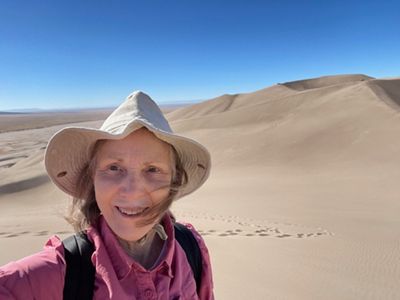 A close-up of a woman wearing a had and pink shirt smiling with a large sand dune behind her.