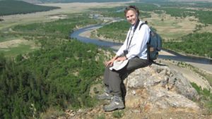 A woman sits on a rock and smiles with a large natural area behind her in the distance.