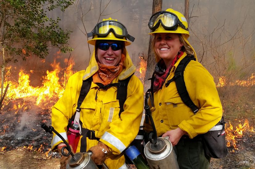 Two women wearing yellow fire retardant gear pose together during a controlled burn. They are both holding silver drip torch canisters. A line of fire burns behind them as smoke rises around them.
