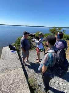Four people stand along the shore of Assawompset Pond on a bright sunny day, recording for the podcast. Two people hold mics, Tom Barron and TNC's Alison Bowden speak into a mic looking at the pond.