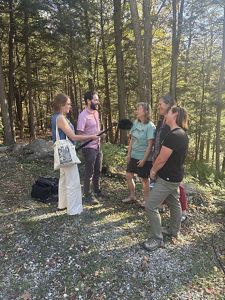 A group of people stand in the clearing of a forest, two holding podcast recording equipment, and three others talking with them.