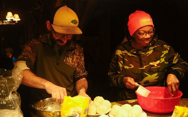 Two people stand smiling looking down at a kitchen counter where they are preparing arepas.