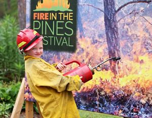 A young person wears a red hard hat and holds a drip torch in front of a festival sign.