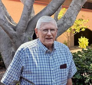Art Smith, an older gentleman with a blue plaid shirt, smiles in front of a large tree on a sunny day.