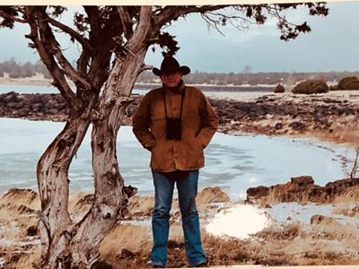 Art Smith, wearing a cowboy hat and binoculars around his neck, stands in front of a small body of water and a tree.