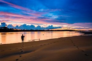 During sunset, a man stands at the shore of the river holding a long stick.