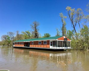 A large barge with a house-like structure on it floats on a flat, calm body of water with trees lining its banks.