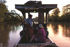 Two people set out on a river on a fishing boat as the sun rises.