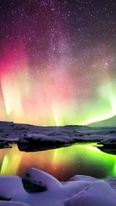 A still pool of water on a snowy night reflects a vibrant aurora borealis and stars from overhead.