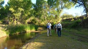 Three individuals with mapping gear walk away from the camera, along a stream of water to their right.