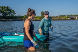 Two women wade through thigh-deep water, pulling a kayak between them.