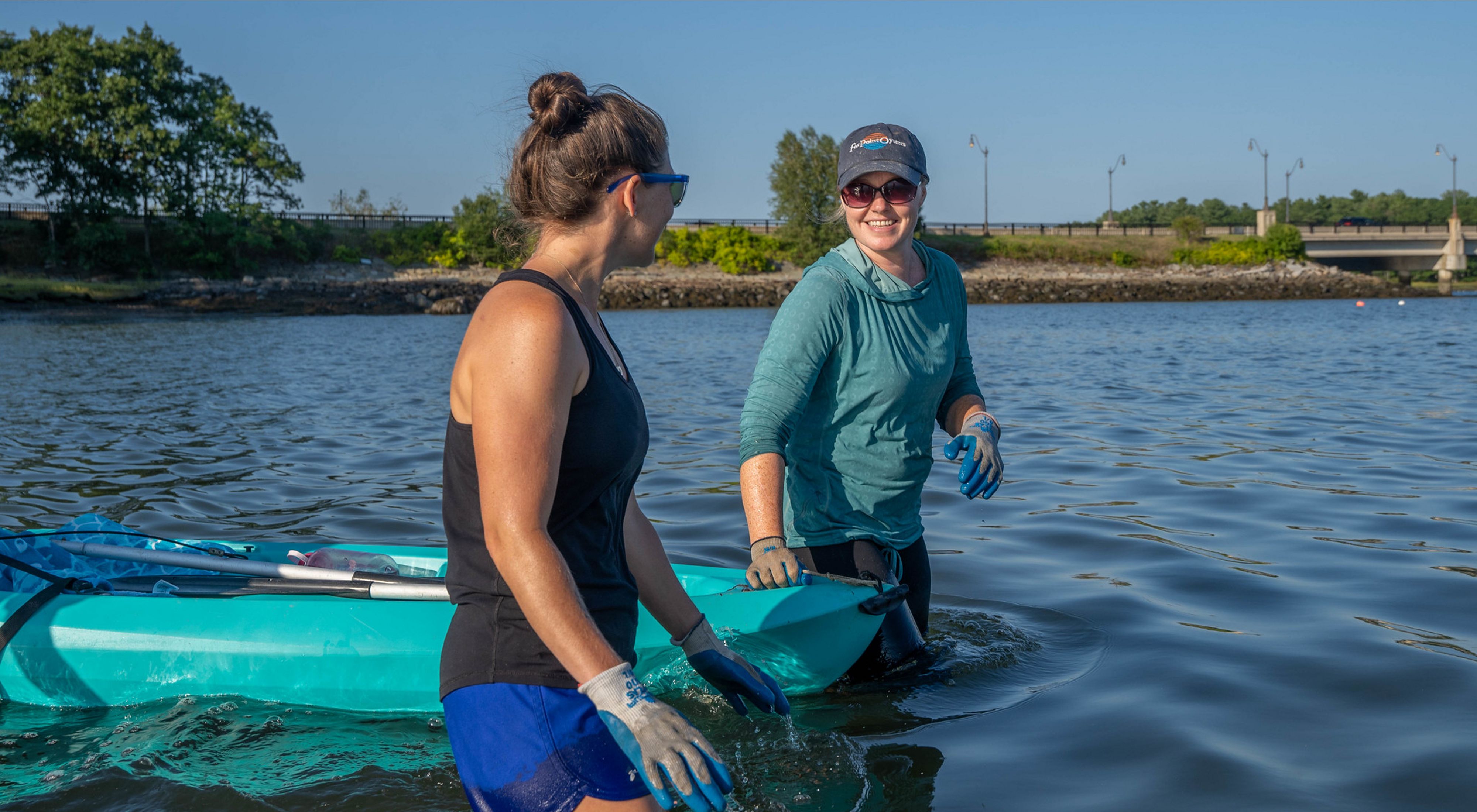 Two women wade through thigh-deep water, pulling a kayak between them.