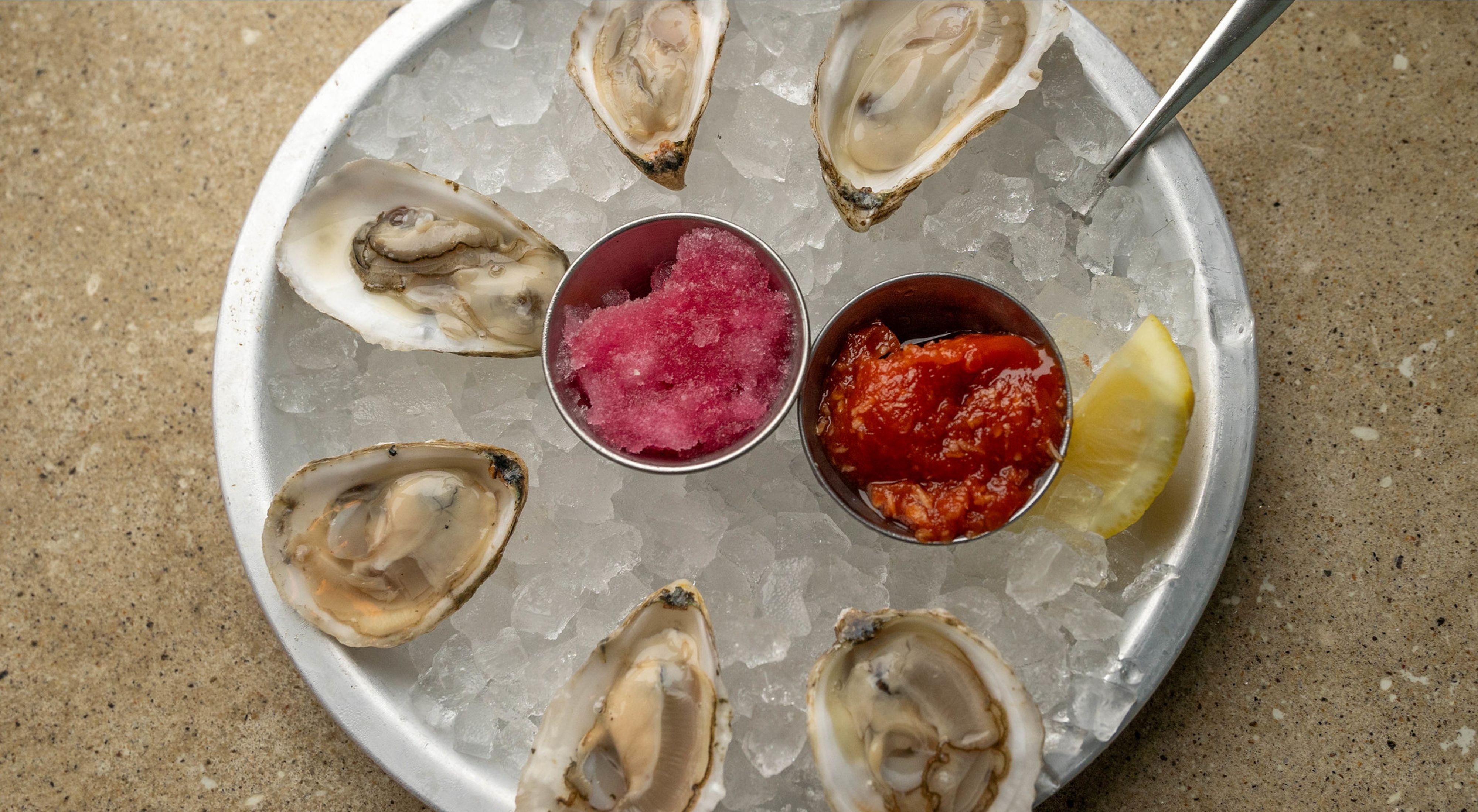 A plate of six oysters on ice, with a lemon wedge and a small container of hot sauce.