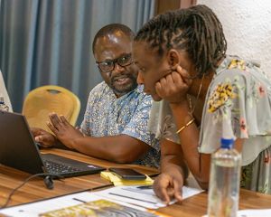 A man sits at a table in front of a laptop. A woman, crouching next to him with her elbows on the table,  is speaking to him.