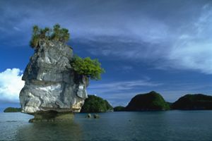 A large rock juts out of the ocean. In the background are small green islands.