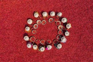 From above, women sit in a circle surrounded by bright red chili peppers. The women all wear woven straw hats and have baskets in front of them.