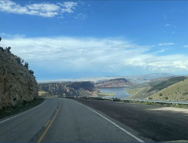 A two-lane highway curves around a bend toward distant mesas. A large lage is visible in the valley below.