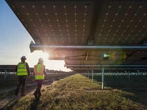 Two workers in safety vests and helmets walk next to solar panels. 