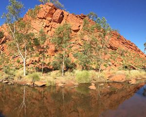 A large hill made of red rocks stands next to a placid stream.