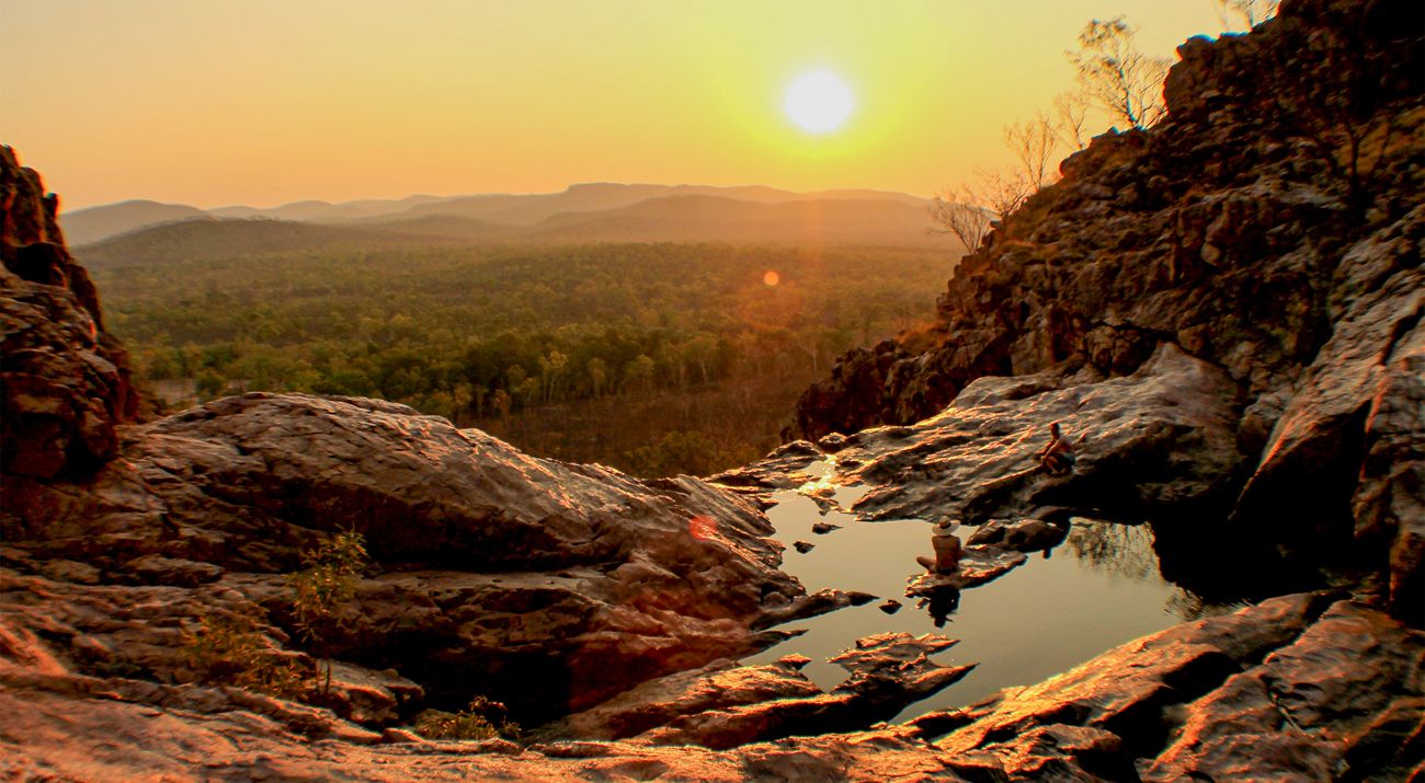 Two people sitting on the edge of a rock pool overlooking bushland at sunset