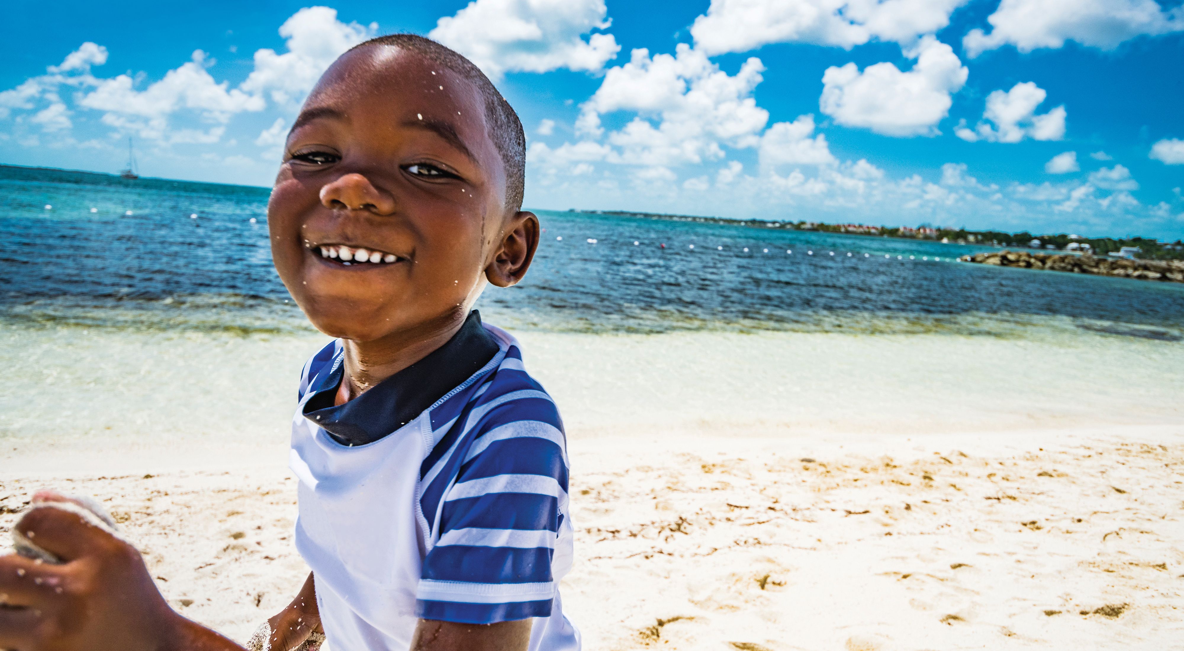 A young boy smiles as he runs along a sandy beach, with blue water and sky behind him.