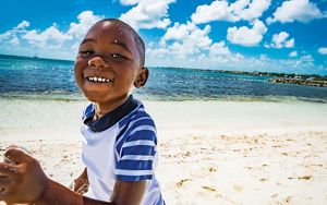 A young boy smiles as he runs along a sandy beach, with glistening ocean and blue sky beyond.