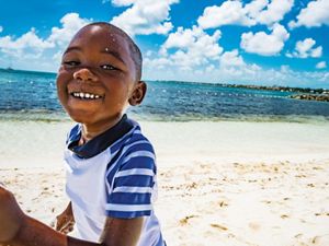 A young boy smiles as he runs along a sandy beach, with glistening ocean and blue sky beyond.