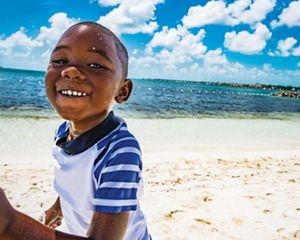 A young boy smiles as he runs along a sandy beach, with glistening ocean and blue sky beyond.