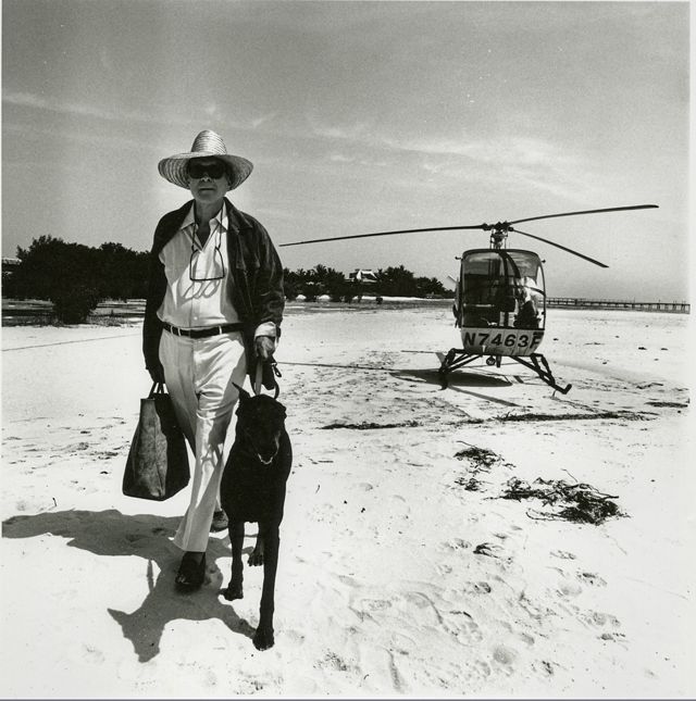black and white image of stylish man with dog walking on sand away from a helicopter