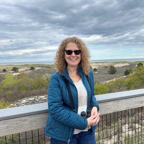 Headshot of Barbara Charry wearing sunglasses and a blue jackert with the beach in the background.