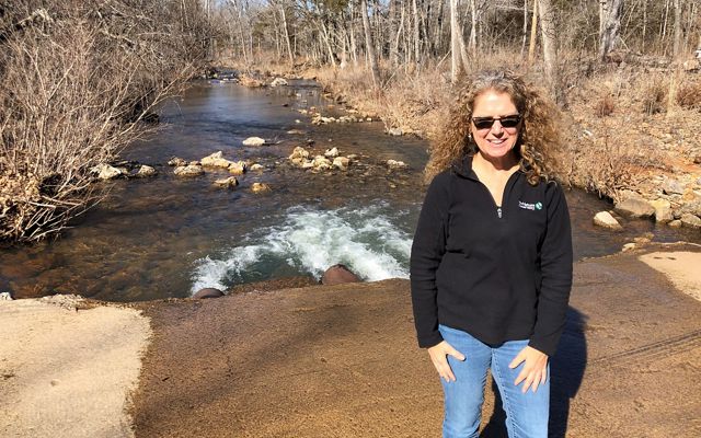 Barbara Charry stands on a concrete dam over Little Shoal Creek with the creek, surrounded by bare winter trees, extending behind her.