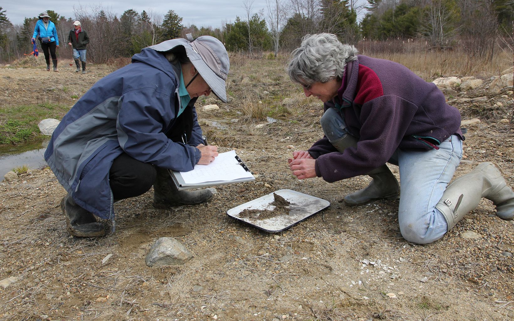 Two women kneel over a tray of soil. One of them digs through while the other takes notes.