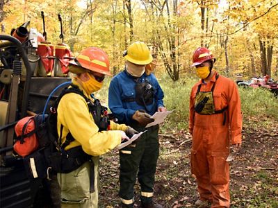 Three people dressed in protecting clothing and gear gather at the back of an UTV and examine written plans.